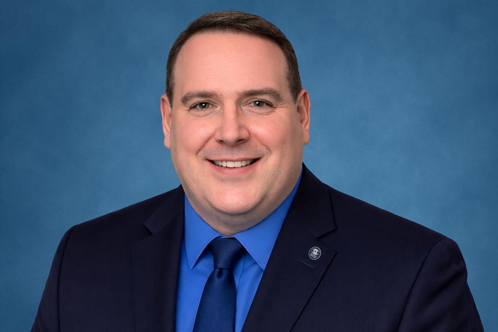Dennis Zehner in a black suit with a blue shirt and tie and Sights for Hope pin smiling against a solid blue background