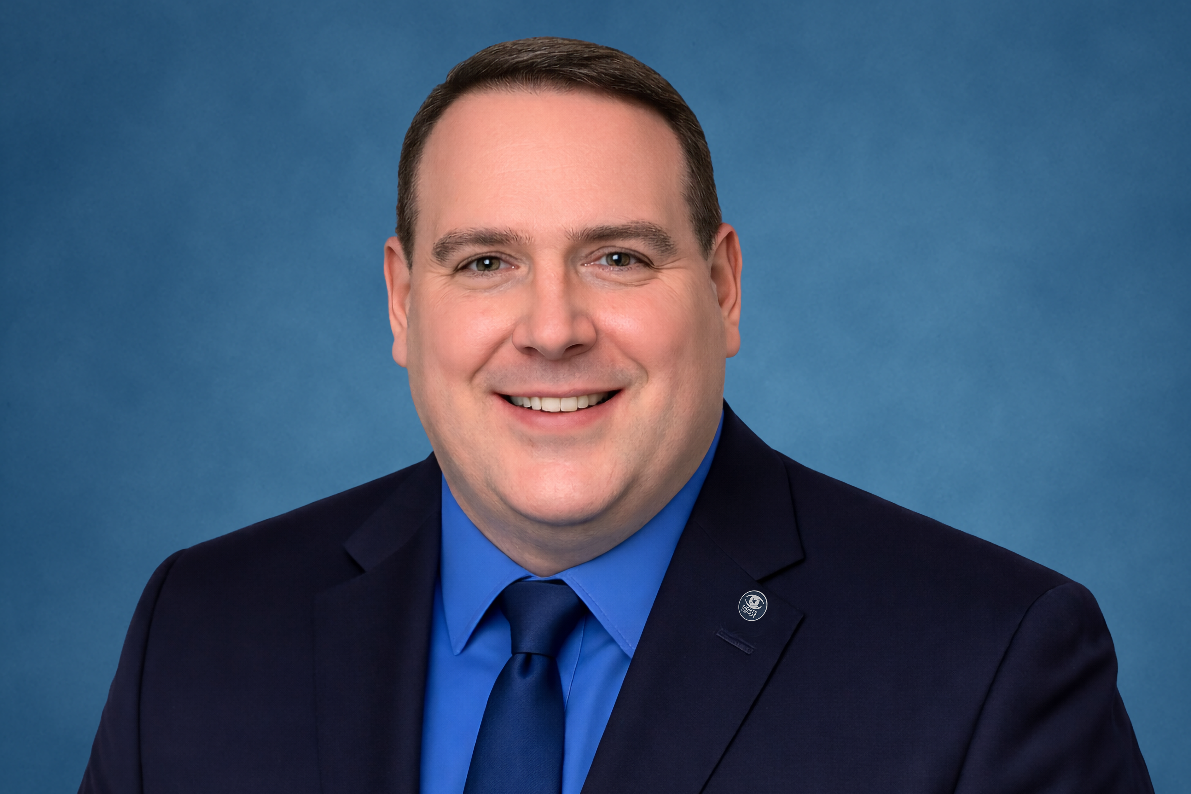 Dennis Zehner in a black suit with a blue shirt and tie and a Sights for Hope lapel pin smiling against a solid blue background.