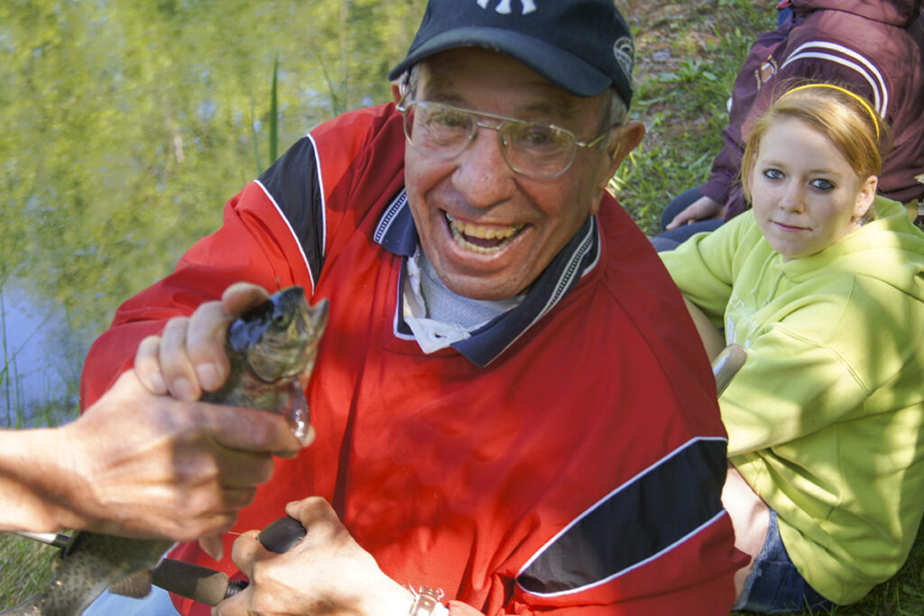 An older man in a red jacket, smiling broadly, holding a small fish near a pond next to a young girl in a lime sweatshirt.