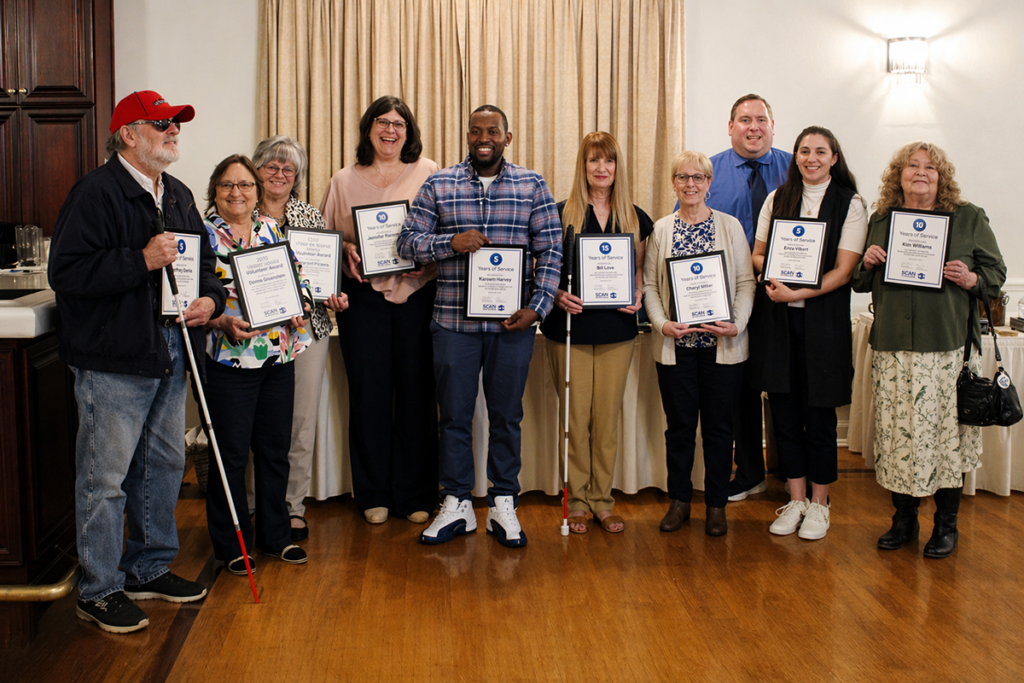 Ten adults standing and smiling in a banquet room, with nine of them holding framed awards recognizing years of service and volunteer excellence.