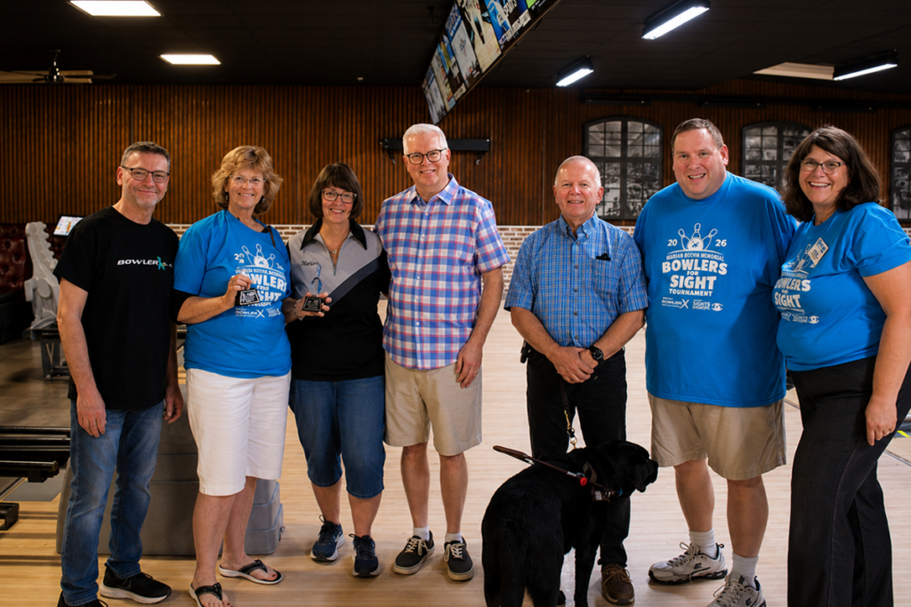 Tournament organizers, sponsors, volunteers, and participants gather for a group photo on the bowling lanes during the 2026 Marian Boehm Memorial Bowlers for Sight Tournament. A guide dog sits in the foreground.