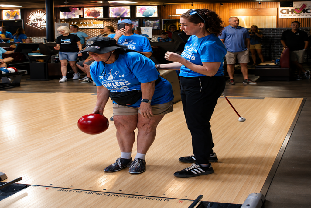 A Sights for Hope client prepares to bowl with assistance from a volunteer during the Marian Boehm Memorial Bowlers for Sight Tournament as participants and spectators look on in the background.