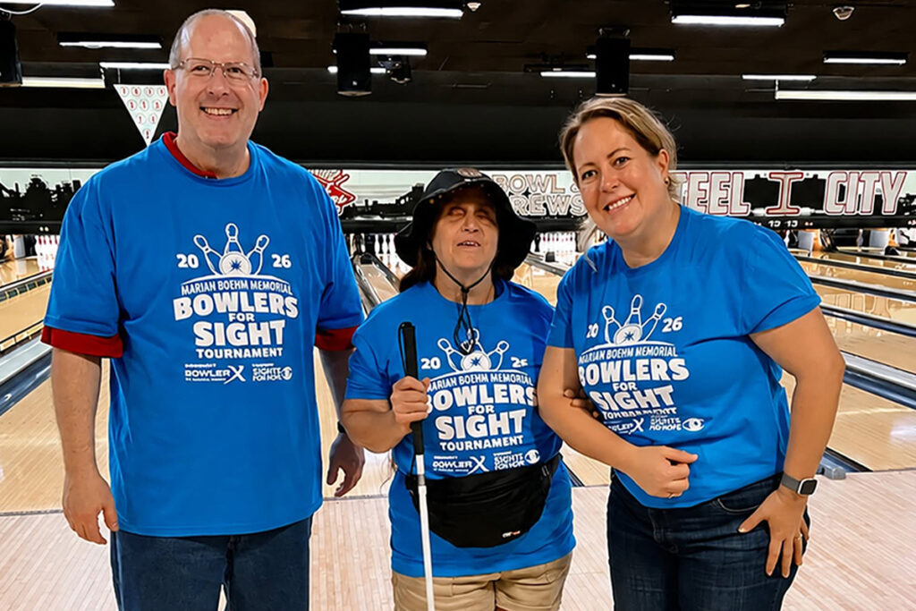 Three participants standing together on a bowling lane during the 2026 Marian Boehm Memorial Bowlers for Sight Tournament. The individuals wear matching blue event shirts, including a participant who is blind holding a white cane, with bowling lanes visible in the background.