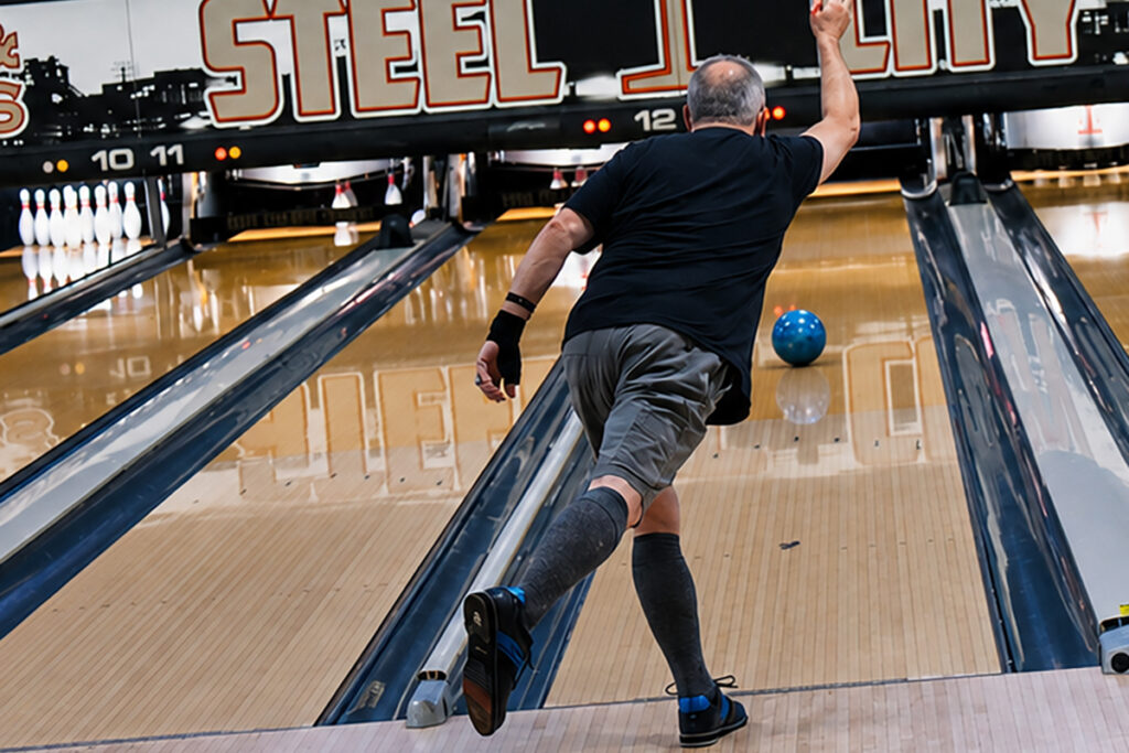 A bowler releasing a blue bowling ball down a lane at Steel City Bowl & Brews. The image is taken from behind, capturing the bowler’s follow-through as pins await at the far end of the lane.