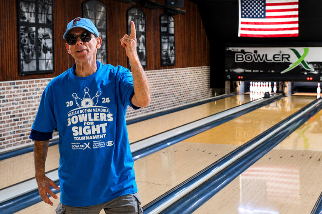 A participant wearing sunglasses and a blue Marian Boehm Memorial Bowlers for Sight Tournament shirt while standing beside a bowling lane. The individual raises one finger in celebration, with bowling lanes, an American flag, and BowlerX signage visible in the background.