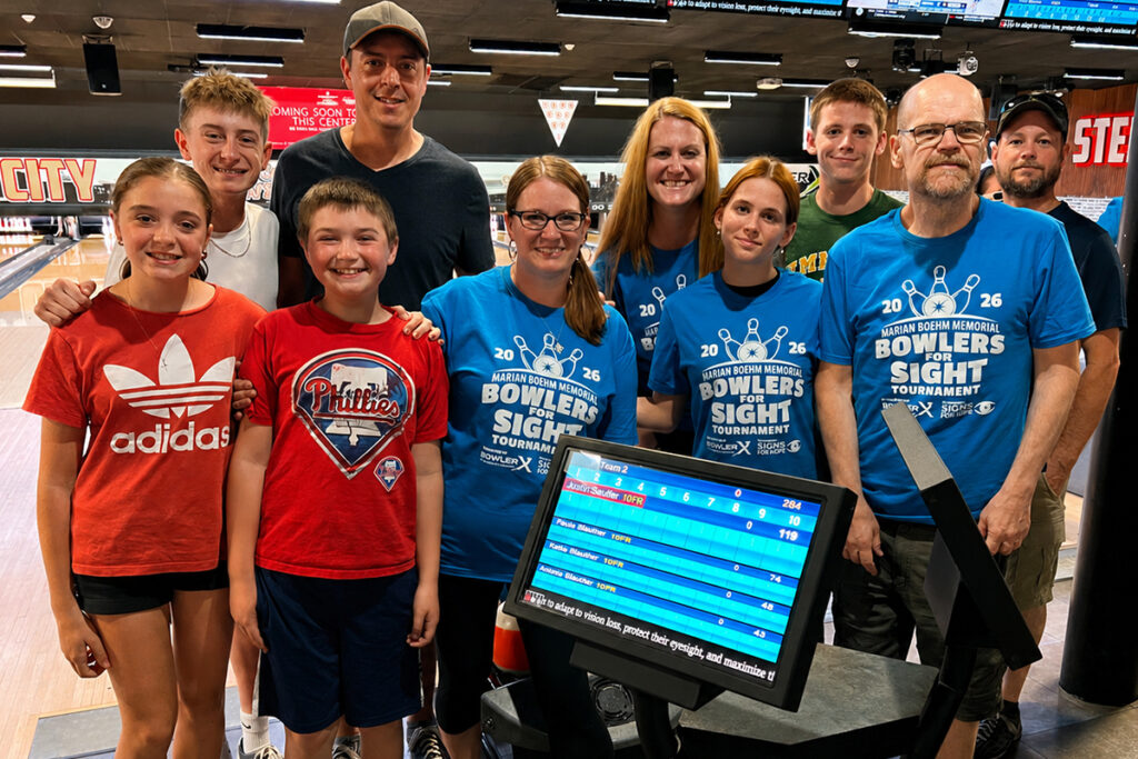 Participants of various ages, including a Sights for Hope client bowler, posing as a group during the inaugural Marian Boehm Memorial Bowlers for Sight Tournament at Steel City Bowl & Brews with bowling lanes and scoreboards visible in the background.