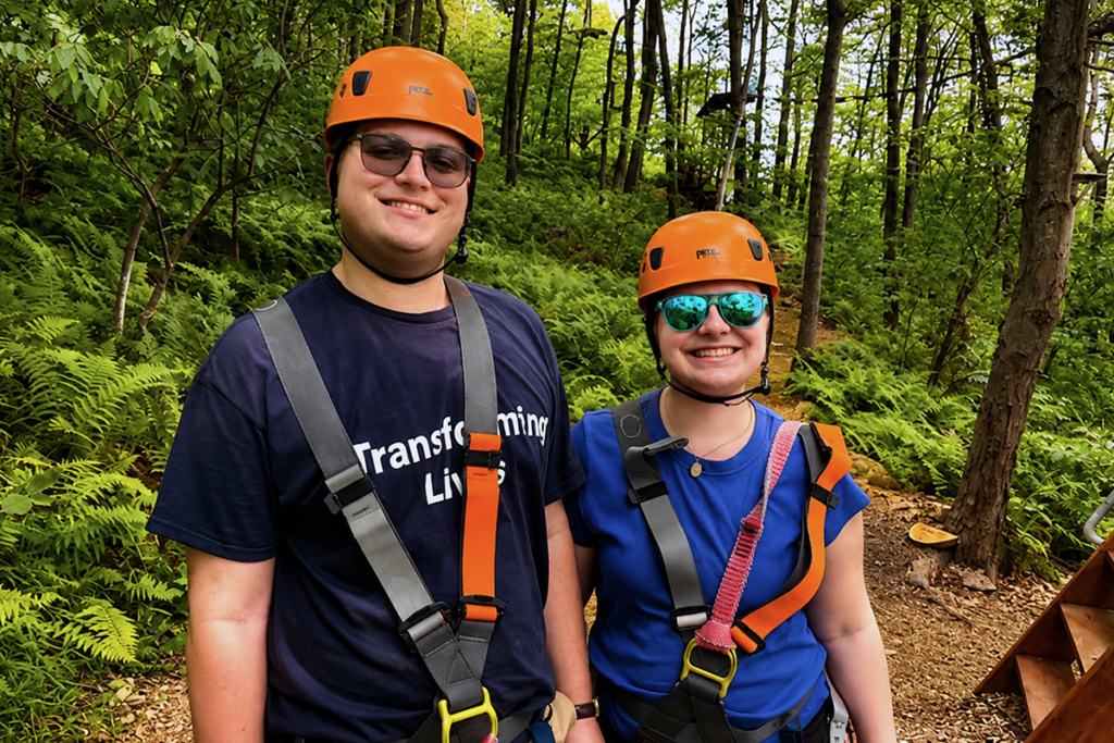 Two participants in Sights for Hope's Transitions program for teens and pre-teens stand by an outdoor ropes course on a wooded trail wearing orange safety helmets and full-body climbing harnesses.