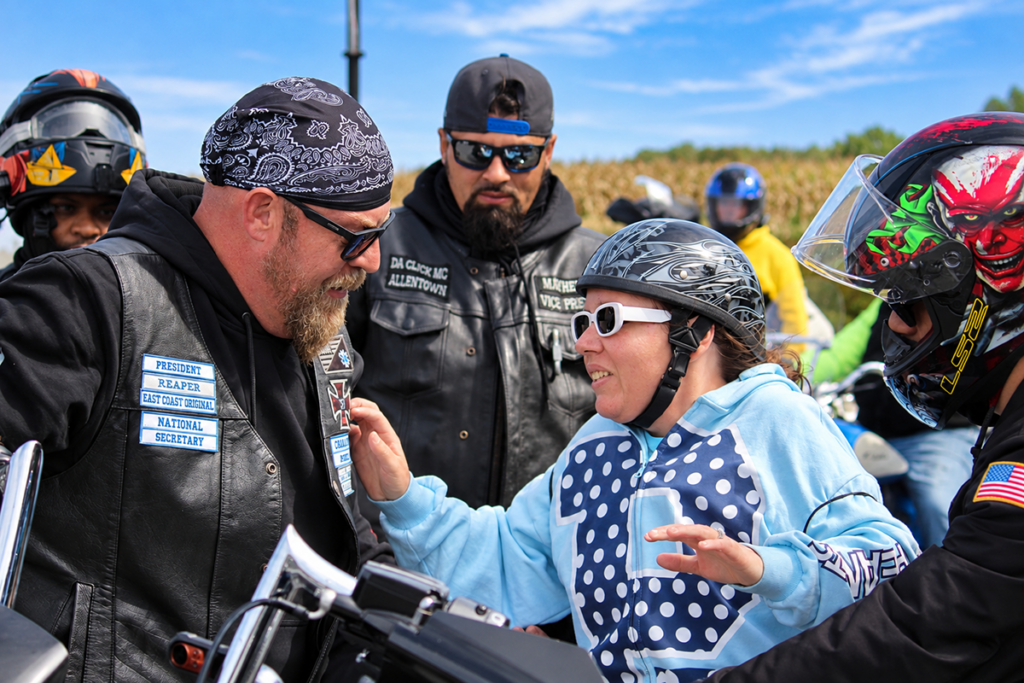 A Sights for Hope client wearing white sunglasses, a patterned motorcycle helmet, and a blue hoodie smiles while talking with two bikers wearing black leather vests during the 2026 Bikers for Sight Ride.