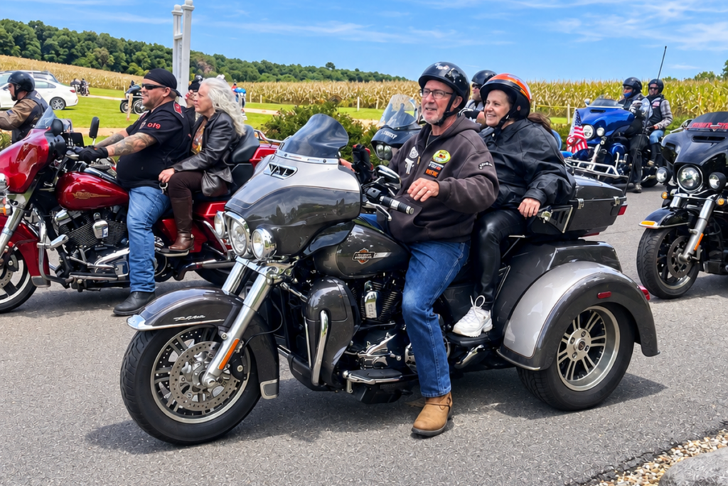 A motorcyclist wearing a black helmet and brown jacket sits on a gray Harley-Davidson trike with a smiling female Sights for Hope client behind him. Other motorcyclists and passengers gather nearby outside the winery.