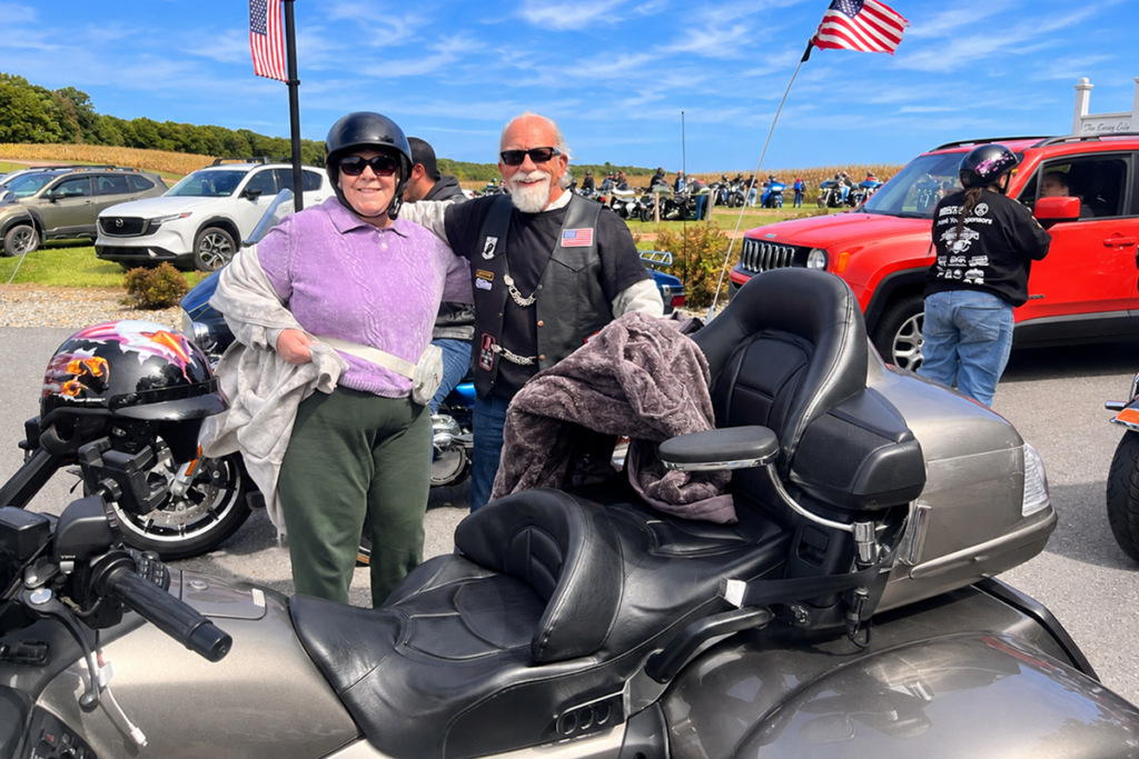 A smiling female Sights for Hope client wearing a purple shirt and motorcycle helmet stands with her arm around a bearded motorcyclist wearing sunglasses and a black vest. They pose beside a large silver touring motorcycle at a winery.