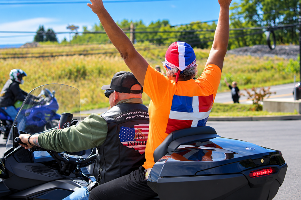A Sights for Hope client wearing a bright orange shirt and a red, white, and blue cap raises both arms high while seated behind a motorcyclist on a black touring motorcycle at the start of the Bikers for Sight Ride.