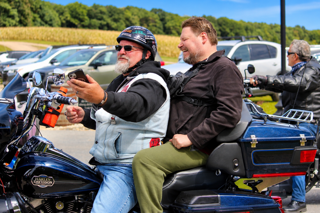 A motorcyclist wearing a helmet, sunglasses, and a denim vest holds up his phone to take a selfie of him and a male Sights for Hope client sitting behind him on a blue Harley-Davidson motorcycle.