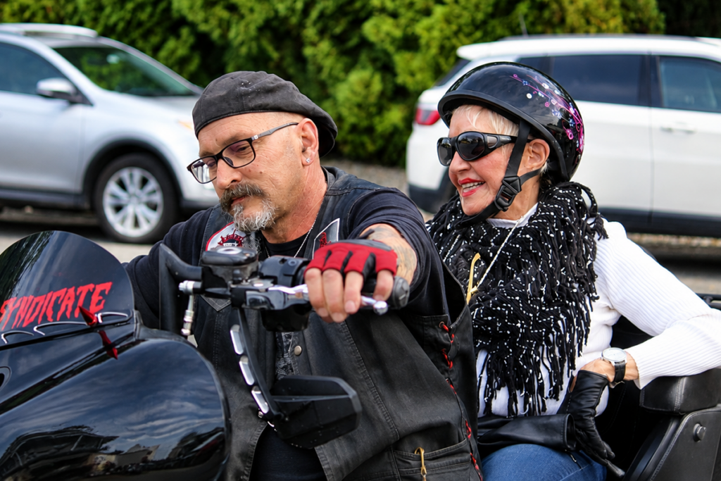 A motorcyclist wearing a black vest and red gloves sits on a black touring motorcycle with a smiling female Sights for Hope client wearing a helmet, sunglasses, and a black-and-white scarf seated behind him.