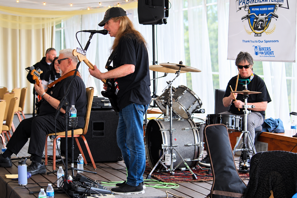 Three members of Limitless perform onstage at the Bikers for Sight Ride. A seated bassist and standing guitarist play alongside a drummer, with a Bikers for Sight sponsor banner hanging behind them.