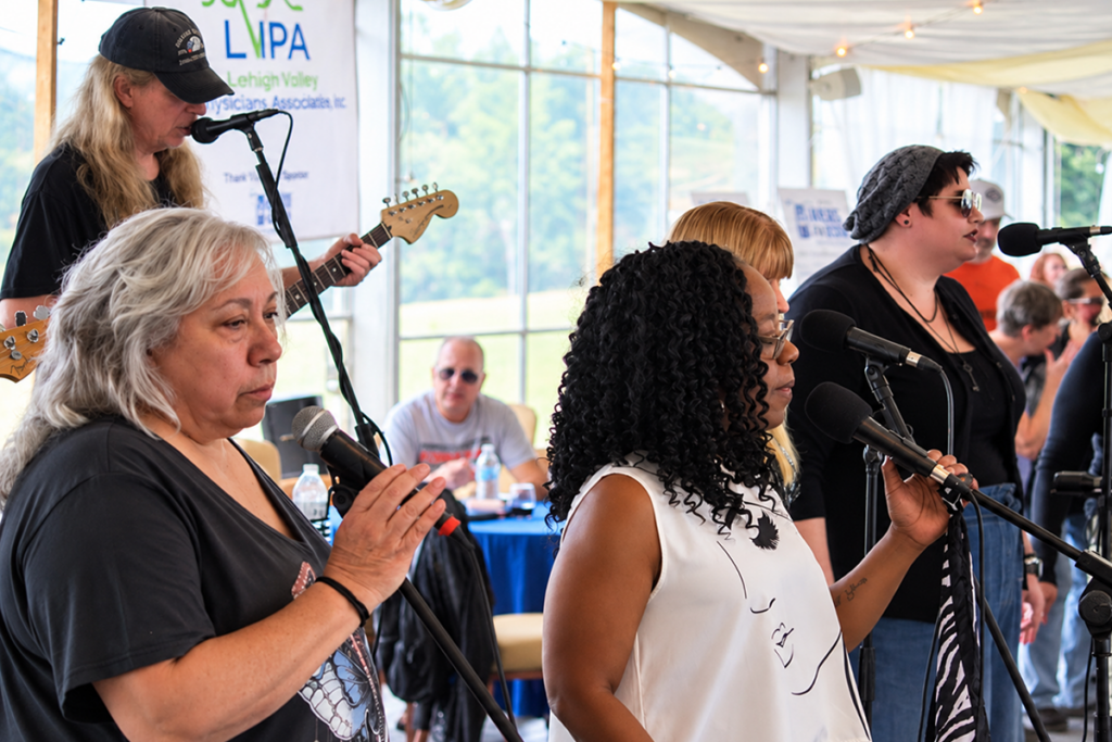 Four vocalists and a guitarist from Limitless perform under a tent at the Bikers for Sight Ride. The women sing into microphones in the foreground while a guitarist plays behind them.