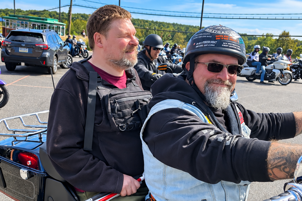 A smiling motorcycle rider wearing a helmet, sunglasses, and a denim vest poses with a Sights for Hope client seated behind him during the 2026 Bikers for Sight Ride.
