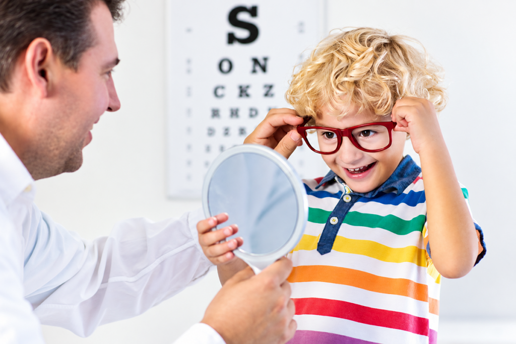A young by smiling as he tries on red eyeglasses and looks at himself in a handheld mirror during an eye care visit.