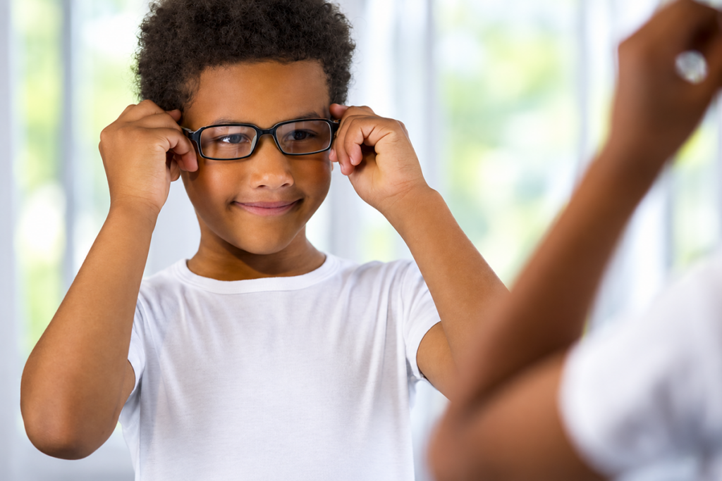 A young boy smiling as he adjusts his black eyeglasses while looking in a mirror.