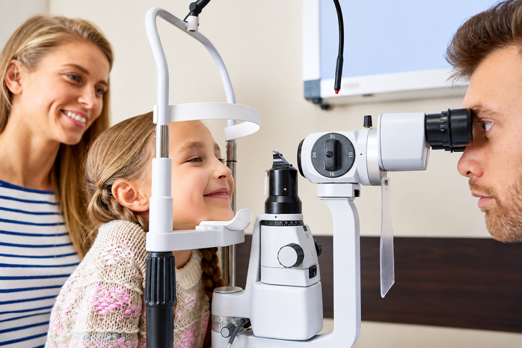 A young girl sitting at a slit lamp while an eye doctor examines her eyes as her mother watches nearby.