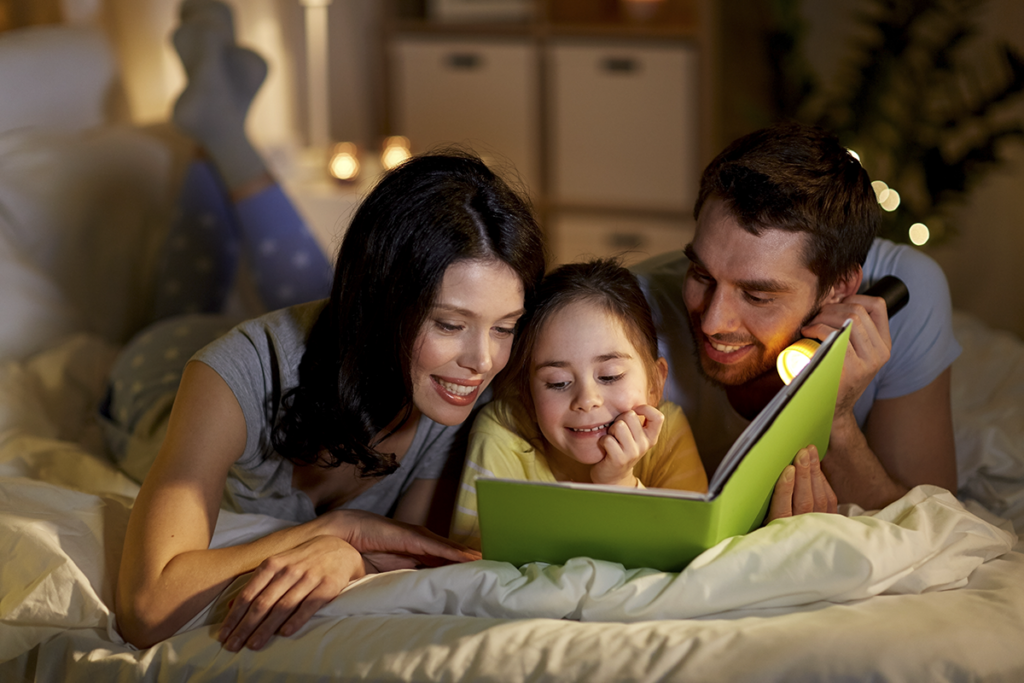 A young girl reading a book in bed with her parents by flashlight in a warmly lit room.