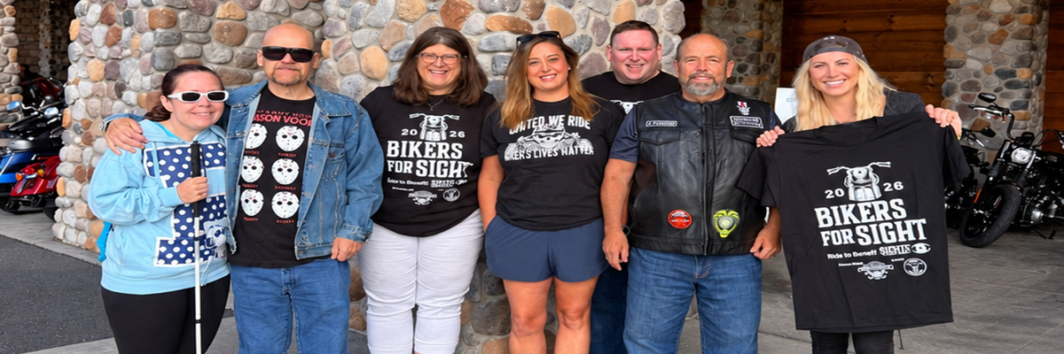 Seven people posing together in front of a stone wall and motorcycles at the 2026 Bikers for Sight event. Several wear Bikers for Sight shirts, one person holds a white cane, and another displays an event shirt.